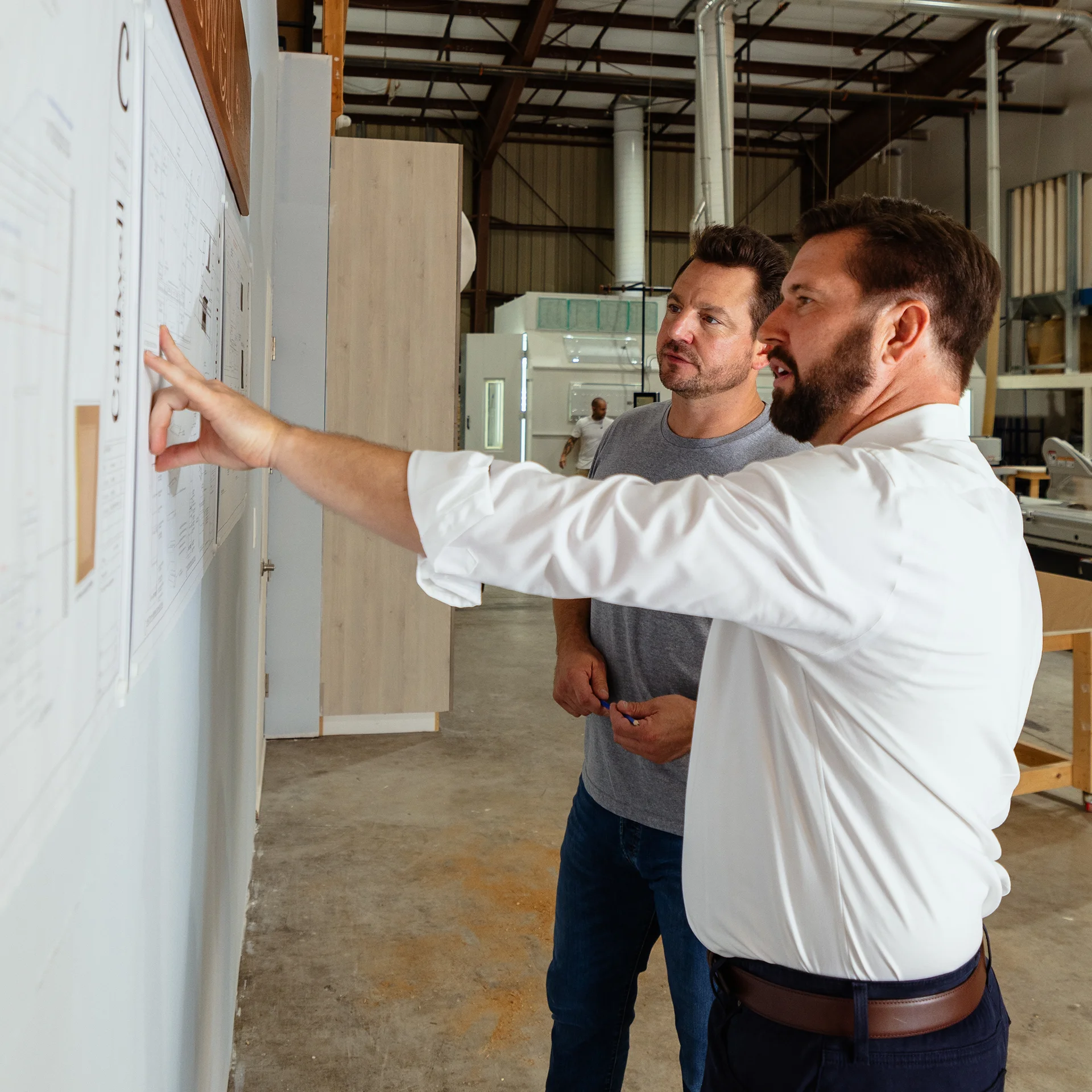Two men in business attire standing before a whiteboard, discussing or explaining blueprints placed on wall.