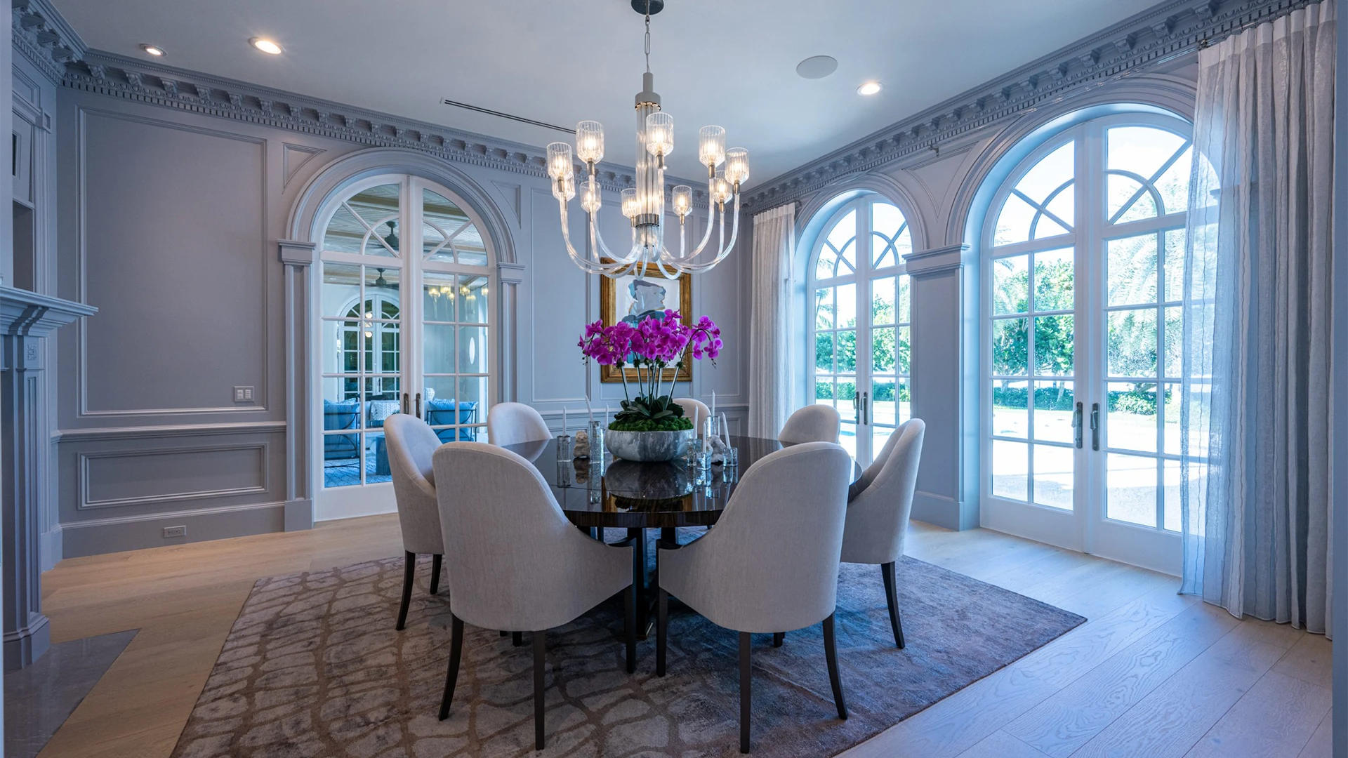 Sophisticated dining room highlighted by a dazzling chandelier, a large white table, and vibrant purple orchids near grand windows.