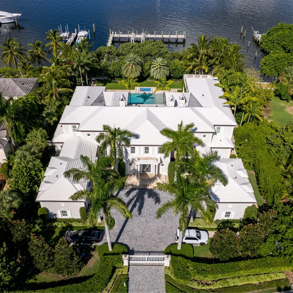 Aerial view of an expansive Bermuda Colonial style home surrounded by lush palm trees and a serene body of water.