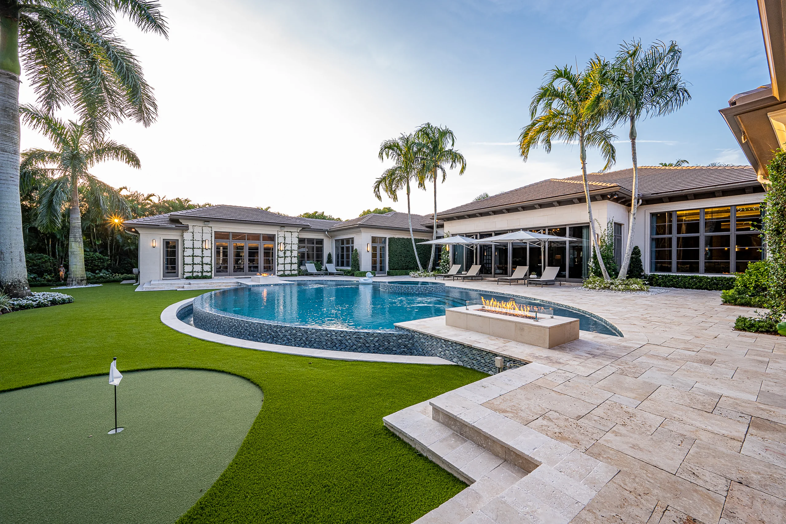 Sunlit backyard featuring a luxurious pool, lush palms, a pristine golf course, and an elegant patio with a cozy fire.